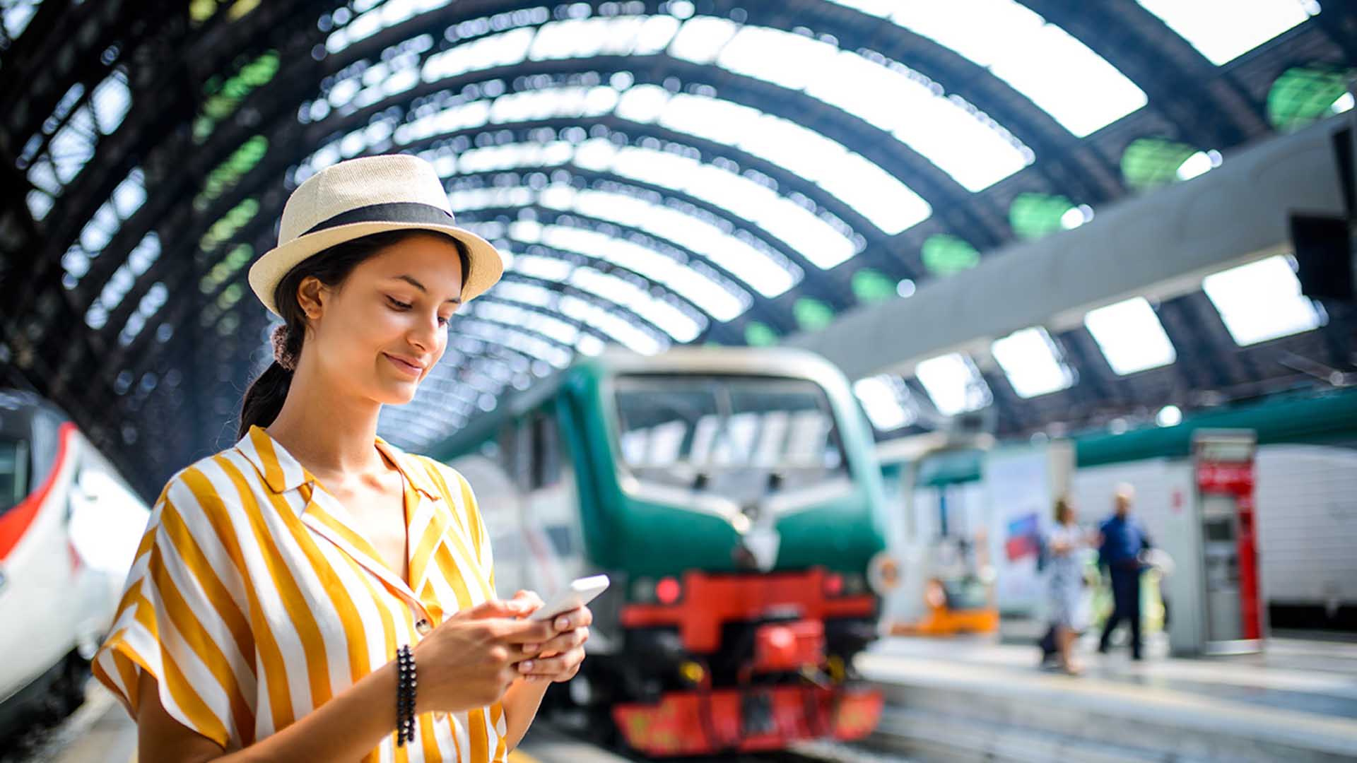 Eine Frau mit Hut schaut am Bahnhof auf ein Telefon
