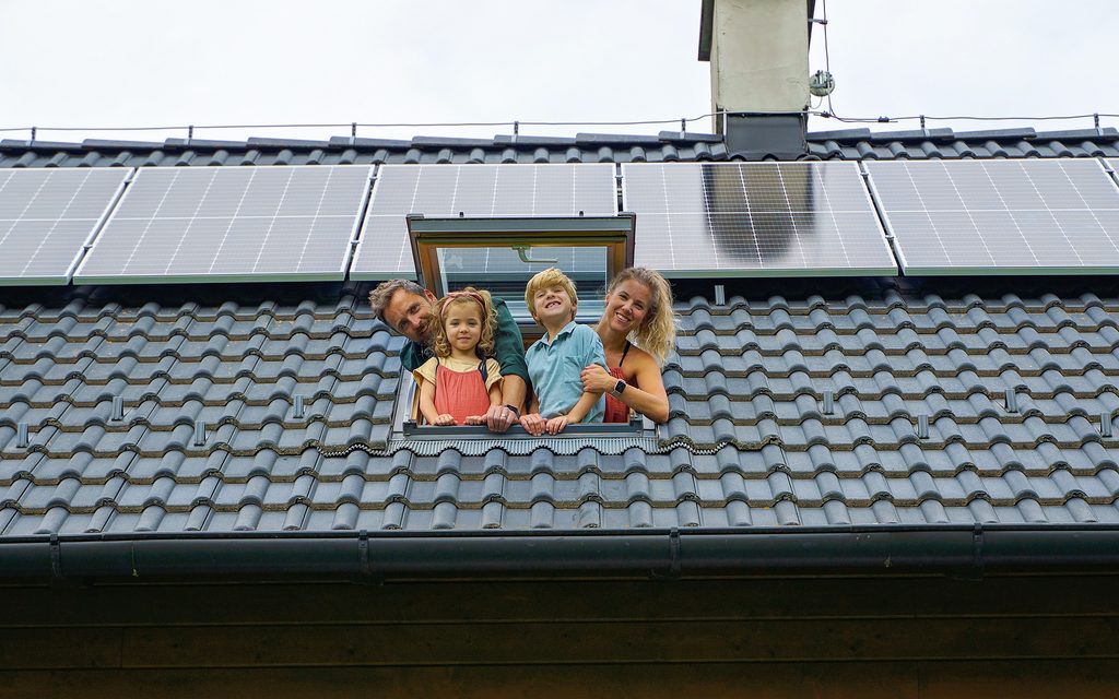 Famiglia felice che si sporge dalla finestra del lucernario nella loro nuova casa con pannelli solari sul tetto. Energia alternativa, risparmio di risorse e concetto di stile di vita sostenibile.